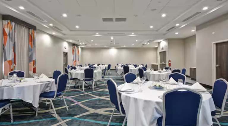 A conference room with round tables covered in white tablecloths, each set with glassware and napkins. Blue chairs surround the tables. The room is well-lit with overhead lights, and abstract pattern carpet covers the floor. Large windows are on the sides.